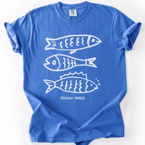 Peut inclure: Un t-shirt bleu à manches courtes avec un motif blanc de trois poissons stylisés et le texte "Piccolo Pesce".