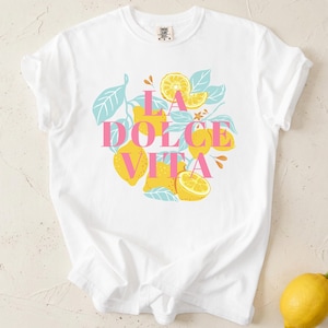 May include: White t-shirt with the text "LA DOLCE VITA" in pink, surrounded by yellow lemons and blue leaves. A single lemon rests on the surface. The shirt is a casual, comfortable style.