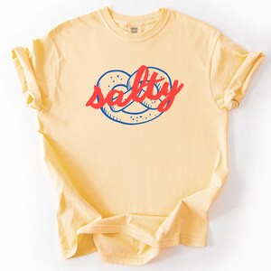 A flat lay of a Butter Comfort Colors shirt with a design featuring a pretzel and the phrase Salty over the top of it.