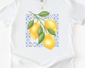 Op de afbeelding: Wit T-shirt met een aquarel illustratie van citroenen op een tak met groene bladeren. De citroenen zijn geplaatst tegen een blauw en wit tegelpatroon. Een zilveren ketting is zichtbaar.