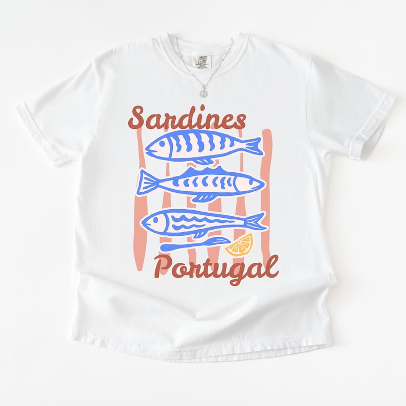 May include: White t-shirt with a graphic design featuring three blue sardines, red vertical stripes, and the text "Sardines Portugal" in brown. A slice of lemon is also included in the design.