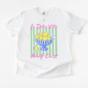 A flat lay of a White Comfort Colors shirt with a. design featuring a bow of lemons against a striped background and the phrase La Dolce Vita, Amalfi Coast in a playful script font.