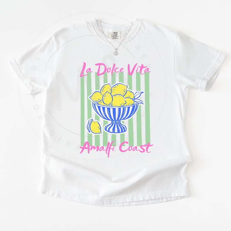 A flat lay of a White Comfort Colors shirt with a. design featuring a bow of lemons against a striped background and the phrase La Dolce Vita, Amalfi Coast in a playful script font.