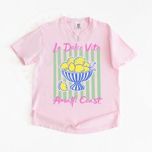 A flat lay of a Blossom Comfort Colors shirt with a. design featuring a bow of lemons against a striped background and the phrase La Dolce Vita, Amalfi Coast in a playful script font.