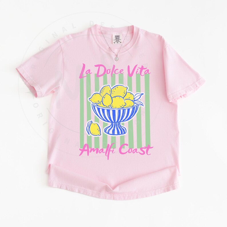 A flat lay of a Blossom Comfort Colors shirt with a. design featuring a bow of lemons against a striped background and the phrase La Dolce Vita, Amalfi Coast in a playful script font.