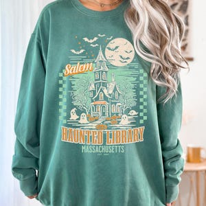 A woman is wearing an oversized Light Green Comfort Colors sweatshirt with a design featuring a haunted library with ghosts and pumpkins and the phrase Salem Haunted Library, Massachusetts.