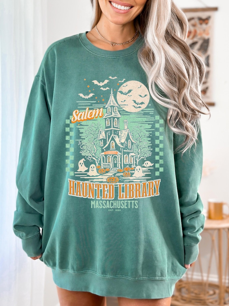 A woman is wearing an oversized Light Green Comfort Colors sweatshirt with a design featuring a haunted library with ghosts and pumpkins and the phrase Salem Haunted Library, Massachusetts.
