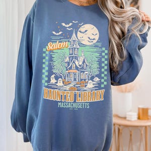 A woman is wearing an oversized Blue Jean Comfort Colors sweatshirt with a design featuring a haunted library with ghosts and pumpkins and the phrase Salem Haunted Library, Massachusetts.