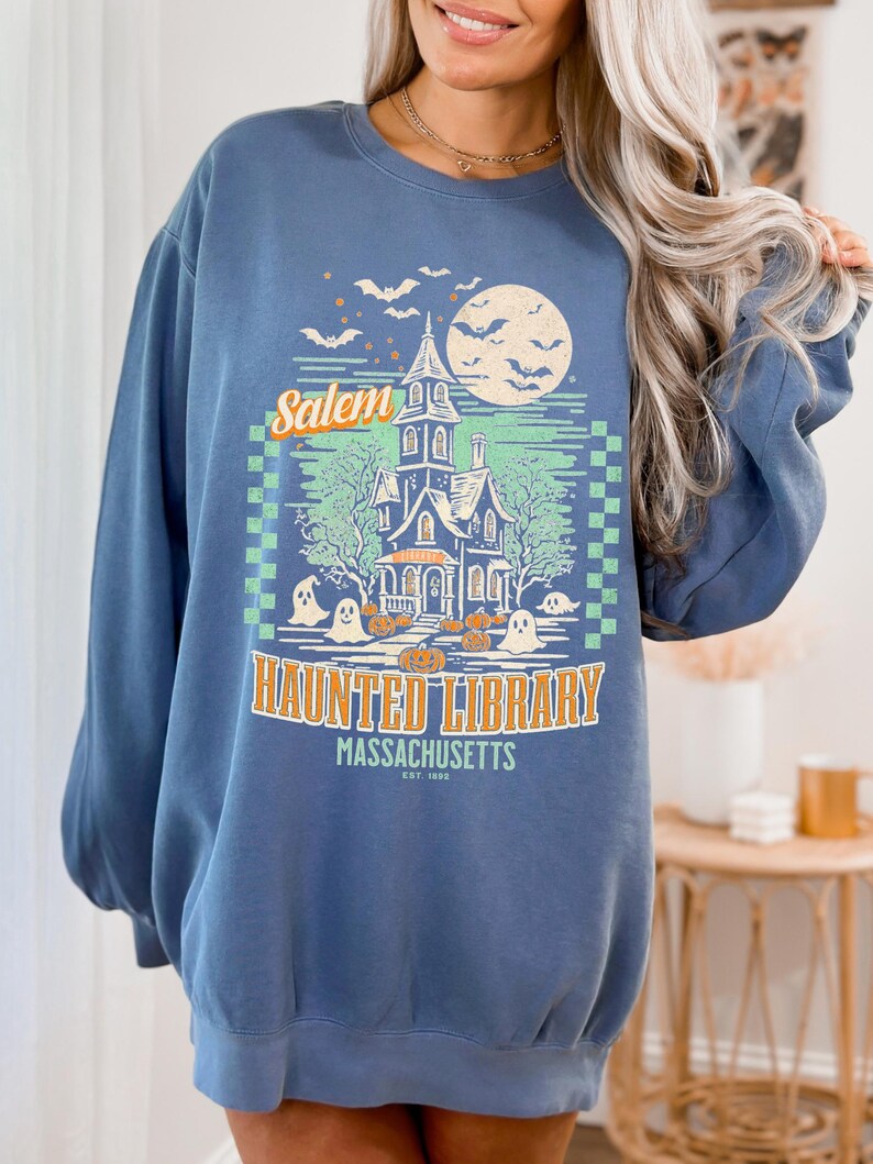 A woman is wearing an oversized Blue Jean Comfort Colors sweatshirt with a design featuring a haunted library with ghosts and pumpkins and the phrase Salem Haunted Library, Massachusetts.