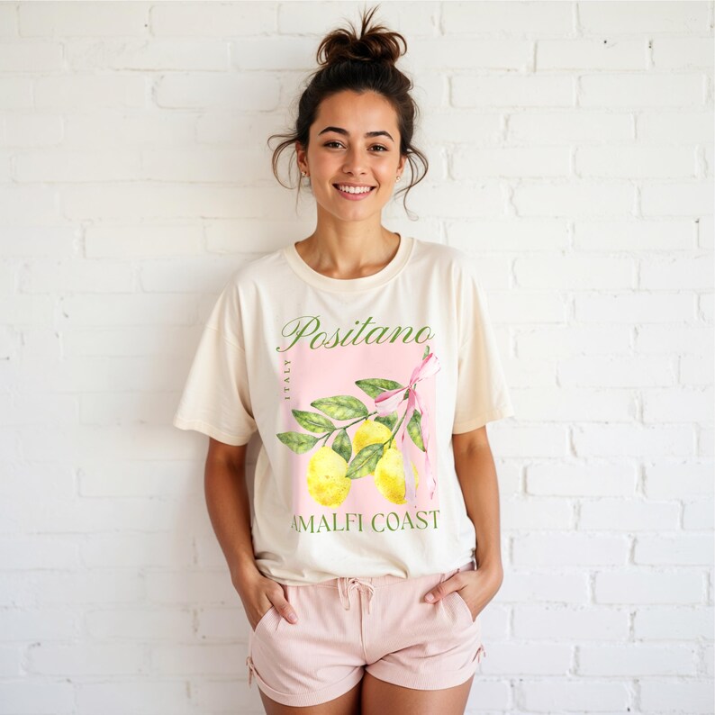 A woman is wearing a Natural Gildan 2000 unisex T-shirt with a design featuring a branch of lemons with a long pink coquette bow tied around it and the phrase Positano, Amalfi Coast, Italy.