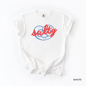 A flat lay of a White Comfort Colors shirt with a design featuring a pretzel and the phrase Salty over the top of it.