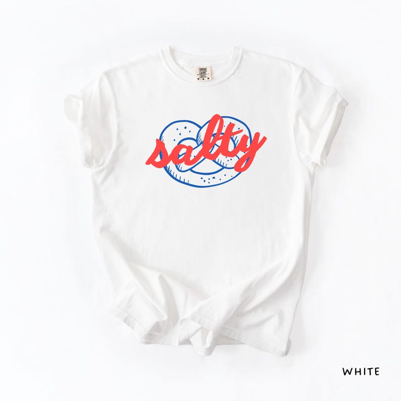 A flat lay of a White Comfort Colors shirt with a design featuring a pretzel and the phrase Salty over the top of it.