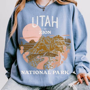 Woman wearing a Blue Jean oversized sweatshirt with a vintage graphic of Zion National Park in Utah, featuring canyons, cacti, and a sunset design, styled with ripped jeans.