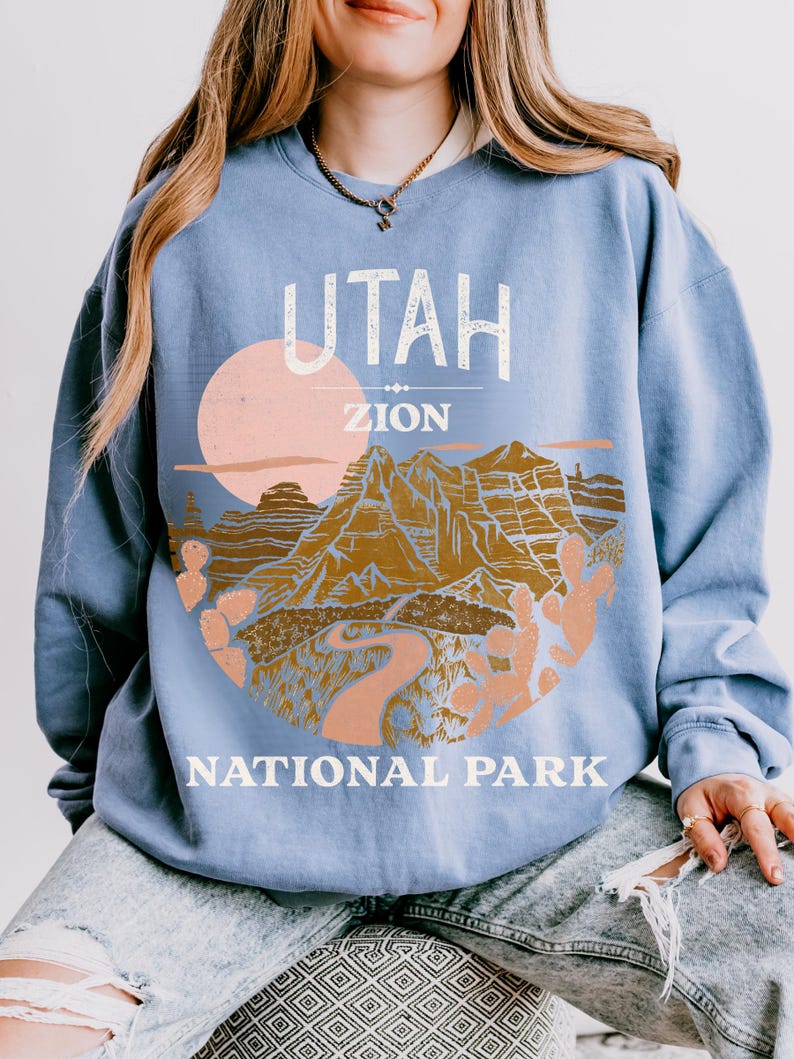 Woman wearing a Blue Jean oversized sweatshirt with a vintage graphic of Zion National Park in Utah, featuring canyons, cacti, and a sunset design, styled with ripped jeans.