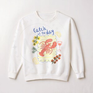 May include: White sweatshirt with a colorful graphic of a lobster, lemon slices, a glass of wine, and the text "Catch of the Day." The design includes yellow and red accents, creating a nautical theme.