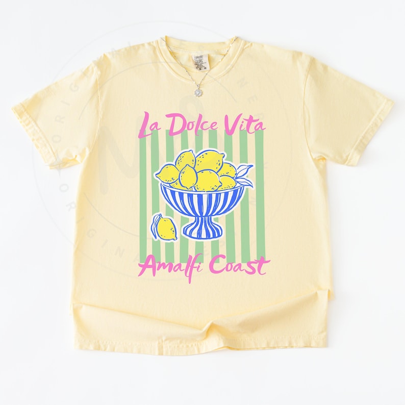 A flat lay of a Banana Comfort Colors shirt with a. design featuring a bow of lemons against a striped background and the phrase La Dolce Vita, Amalfi Coast in a playful script font.