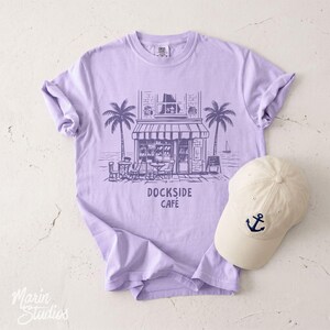 Peut inclure: T-shirt lilas avec un dessin de caf&eacute;, de palmiers et le texte "Dockside Cafe". Une casquette de baseball cr&egrave;me avec un motif d'ancre bleu marine est pos&eacute;e &agrave; c&ocirc;t&eacute;.