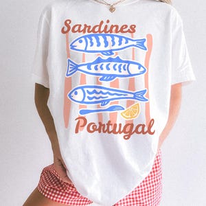 May include: White t-shirt with a graphic design featuring three blue sardines, a lemon slice, and the words "Sardines Portugal" in a retro style. The design is set against a backdrop of vertical peach stripes.