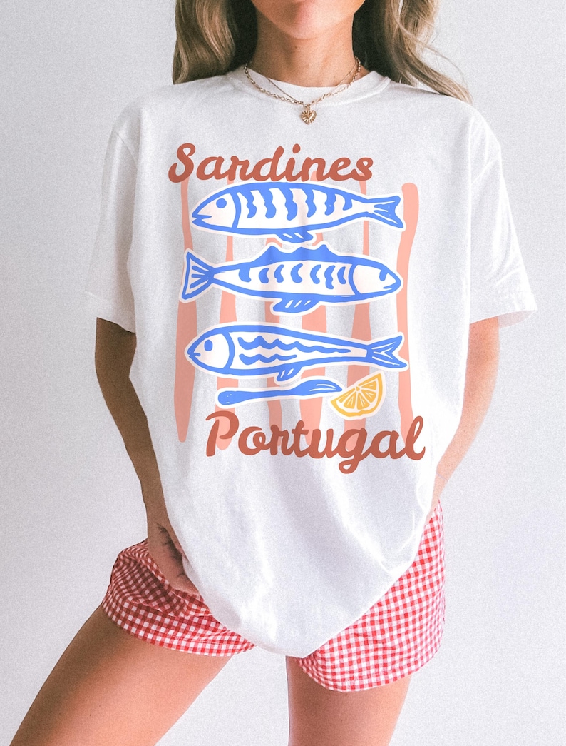 May include: White t-shirt with a graphic design featuring three blue sardines, a lemon slice, and the words "Sardines Portugal" in a retro style. The design is set against a backdrop of vertical peach stripes.