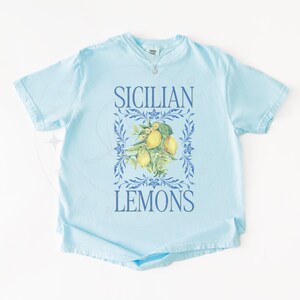 May include: Light blue t-shirt with the words "SICILIAN LEMONS" in blue. The shirt features a floral design with lemons in the center. The shirt has short sleeves and a crew neck.