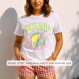 A woman is wearing a white Comfort Colors shirt with a design featuring a hand-drawn lemon and lemon slices on a plate, and the phrase Siciliana at the top in a serif font.