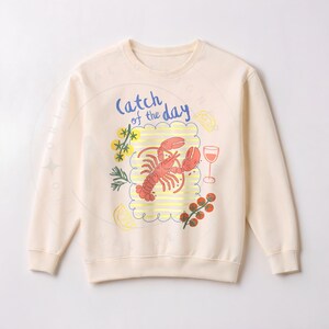 May include: Cream-colored sweatshirt with a colorful graphic of a lobster, wine glass, and other food items. The phrase "Catch of the Day" is written in blue script above the lobster. The design is enclosed in a cloud-like border.