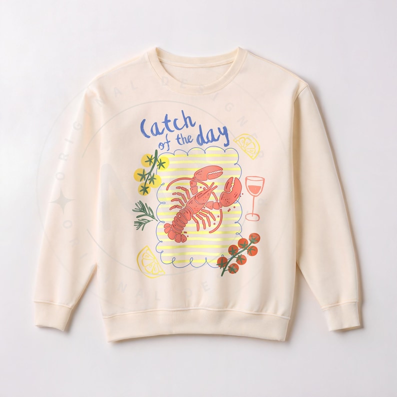 May include: Cream-colored sweatshirt with a colorful graphic of a lobster, wine glass, and other food items. The phrase "Catch of the Day" is written in blue script above the lobster. The design is enclosed in a cloud-like border.