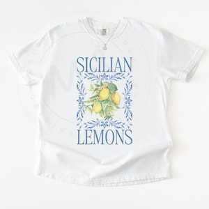 May include: White t-shirt with a blue and yellow graphic. The graphic features the words "SICILIAN LEMONS" in blue, framing a cluster of yellow lemons and green leaves. The shirt has a classic crew neck.