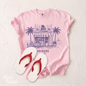 Peut inclure: Un t-shirt rose clair avec un motif de caf&eacute;, des palmiers et le texte "DOCKSIDE COFFEE". Des tongs blanches avec des lani&egrave;res rouges sont au premier plan. Le t-shirt est en mati&egrave;re douce.