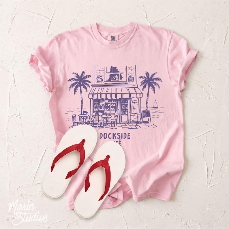 Peut inclure: Un t-shirt rose clair avec un motif de caf&eacute;, des palmiers et le texte "DOCKSIDE COFFEE". Des tongs blanches avec des lani&egrave;res rouges sont au premier plan. Le t-shirt est en mati&egrave;re douce.