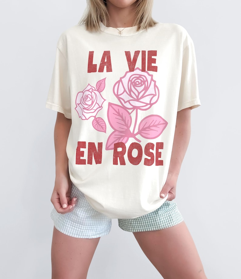 Woman wearing a cream-colored oversized t-shirt with bold red lettering that reads “La Vie En Rose.” The graphic features three outlined pink roses with stylized leaves, giving a vintage-inspired romantic look. She pairs the tee with pastel green gingham shorts for a soft, summery aesthetic.