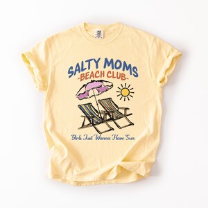 A Butter Comfort Colors shirt ona simple white background with a desgn  featuring a colorful hand-drawn scene of a striped beach umbrella and two chairs with the phrase Salty Moms Beach Club at the top and Girls Just Wanna Have Fun at the bottom of the design.