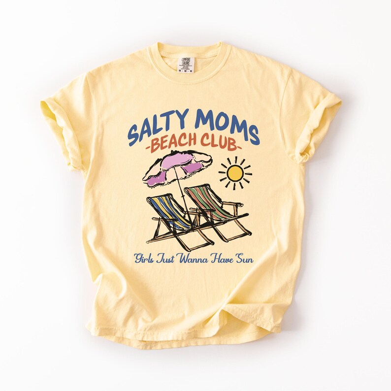 A Butter Comfort Colors shirt ona simple white background with a desgn  featuring a colorful hand-drawn scene of a striped beach umbrella and two chairs with the phrase Salty Moms Beach Club at the top and Girls Just Wanna Have Fun at the bottom of the design.