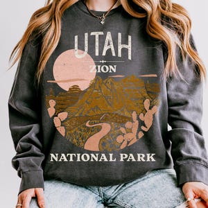 Woman wearing a Pepper oversized sweatshirt with a vintage graphic of Zion National Park in Utah, featuring canyons, cacti, and a sunset design, styled with ripped jeans.