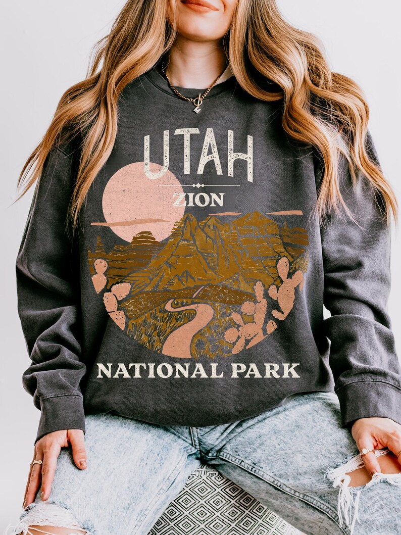 Woman wearing a Pepper oversized sweatshirt with a vintage graphic of Zion National Park in Utah, featuring canyons, cacti, and a sunset design, styled with ripped jeans.