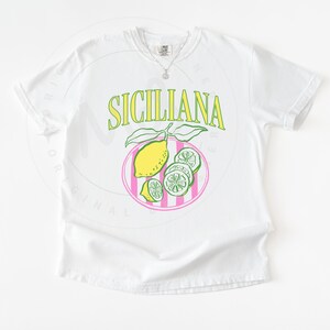 A flat lay of a white Comfort Colors shirt with a design featuring a hand-drawn lemon and lemon slices on a plate, and the phrase Siciliana at the top in a serif font.