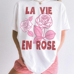 Woman wearing a white oversized t-shirt with bold red lettering that reads “La Vie En Rose.” The graphic features three outlined pink roses with stylized leaves, giving a vintage-inspired romantic look. She pairs the tee with pastel green gingham shorts for a soft, summery aesthetic.