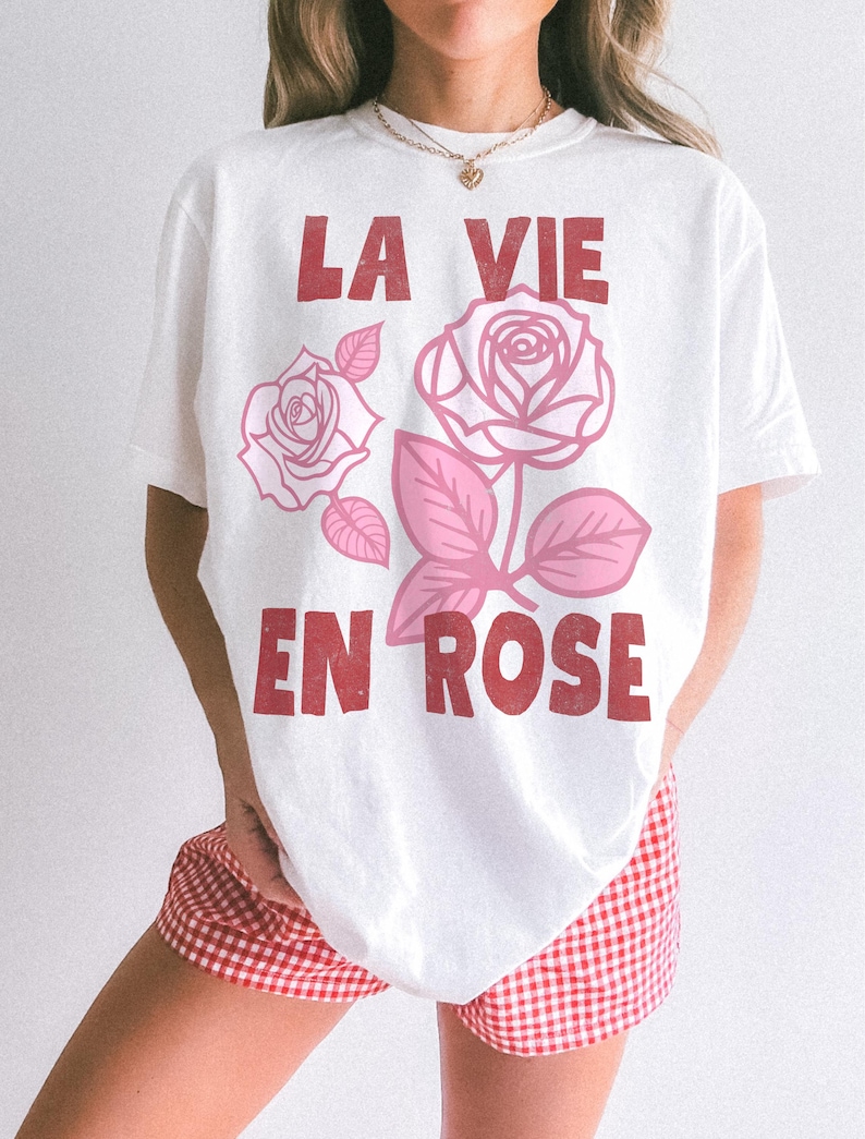 Woman wearing a white oversized t-shirt with bold red lettering that reads “La Vie En Rose.” The graphic features three outlined pink roses with stylized leaves, giving a vintage-inspired romantic look. She pairs the tee with pastel green gingham shorts for a soft, summery aesthetic.