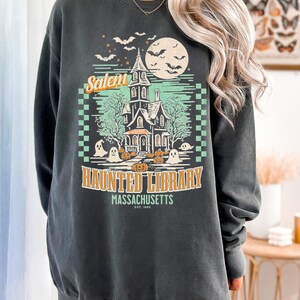 A woman is wearing an oversized Pepper Comfort Colors sweatshirt with a design featuring a haunted library with ghosts and pumpkins and the phrase Salem Haunted Library, Massachusetts.