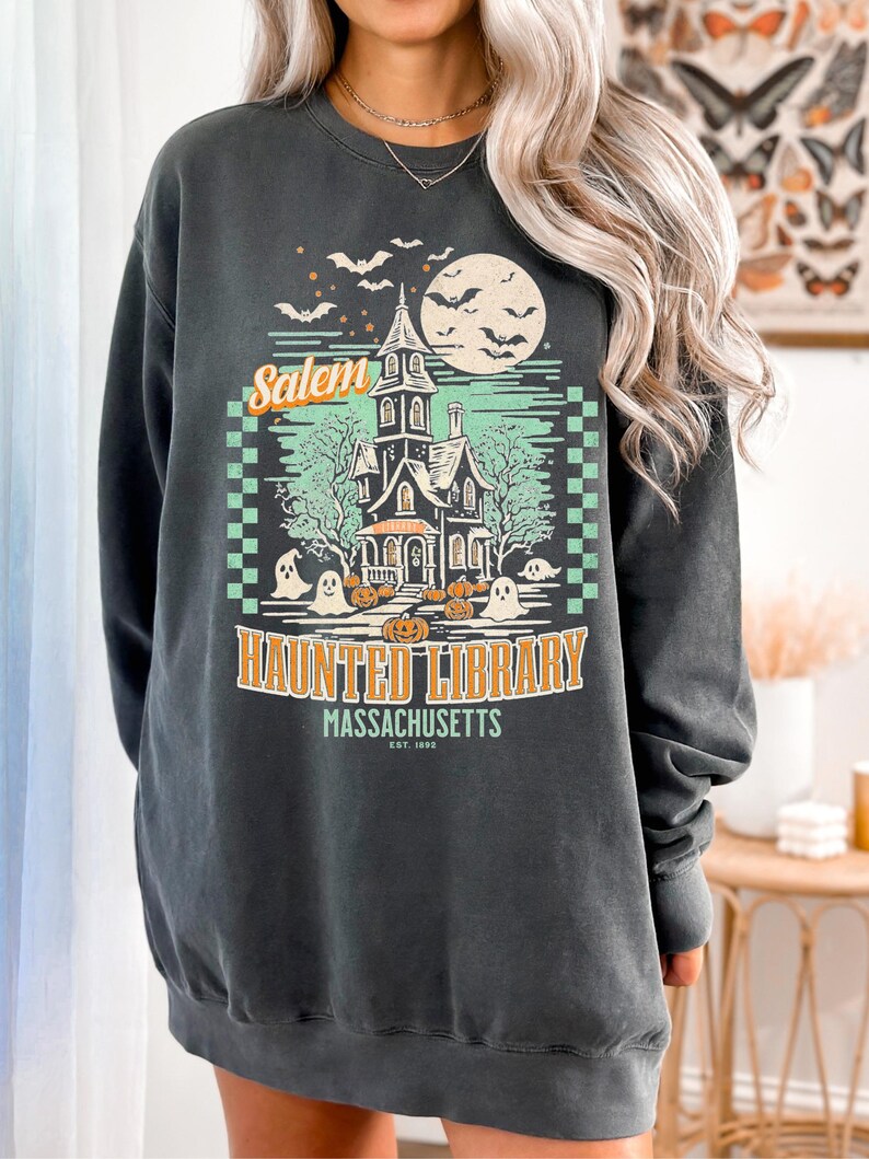 A woman is wearing an oversized Pepper Comfort Colors sweatshirt with a design featuring a haunted library with ghosts and pumpkins and the phrase Salem Haunted Library, Massachusetts.