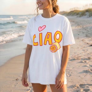 K&ouml;nnte beinhalten: Wei&szlig;es T-Shirt mit dem Wort "CIAO" in Gelb und Orange, einem rosa Herz und einer Zitronen-Grafik. Das T-Shirt wird an einem Strand mit dem Meer im Hintergrund getragen.