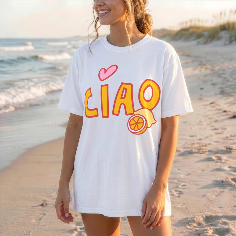 K&ouml;nnte beinhalten: Wei&szlig;es T-Shirt mit dem Wort "CIAO" in Gelb und Orange, einem rosa Herz und einer Zitronen-Grafik. Das T-Shirt wird an einem Strand mit dem Meer im Hintergrund getragen.