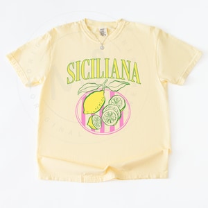 A flat lay of a banana Comfort Colors shirt with a design featuring a hand-drawn lemon and lemon slices on a plate, and the phrase Siciliana at the top in a serif font.