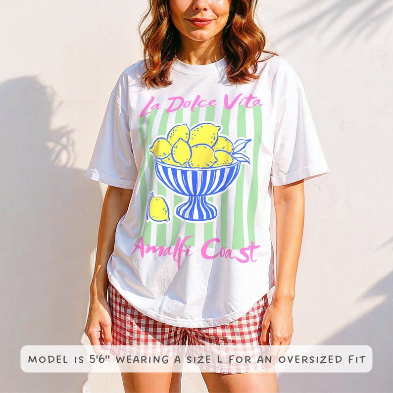 A woman is wearing a White Comfort Colors shirt with a. design featuring a bow of lemons against a striped background and the phrase La Dolce Vita, Amalfi Coast in a playful script font.