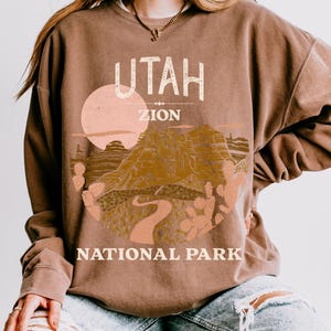 Woman wearing an Espresso oversized sweatshirt with a vintage graphic of Zion National Park in Utah, featuring canyons, cacti, and a sunset design, styled with ripped jeans.