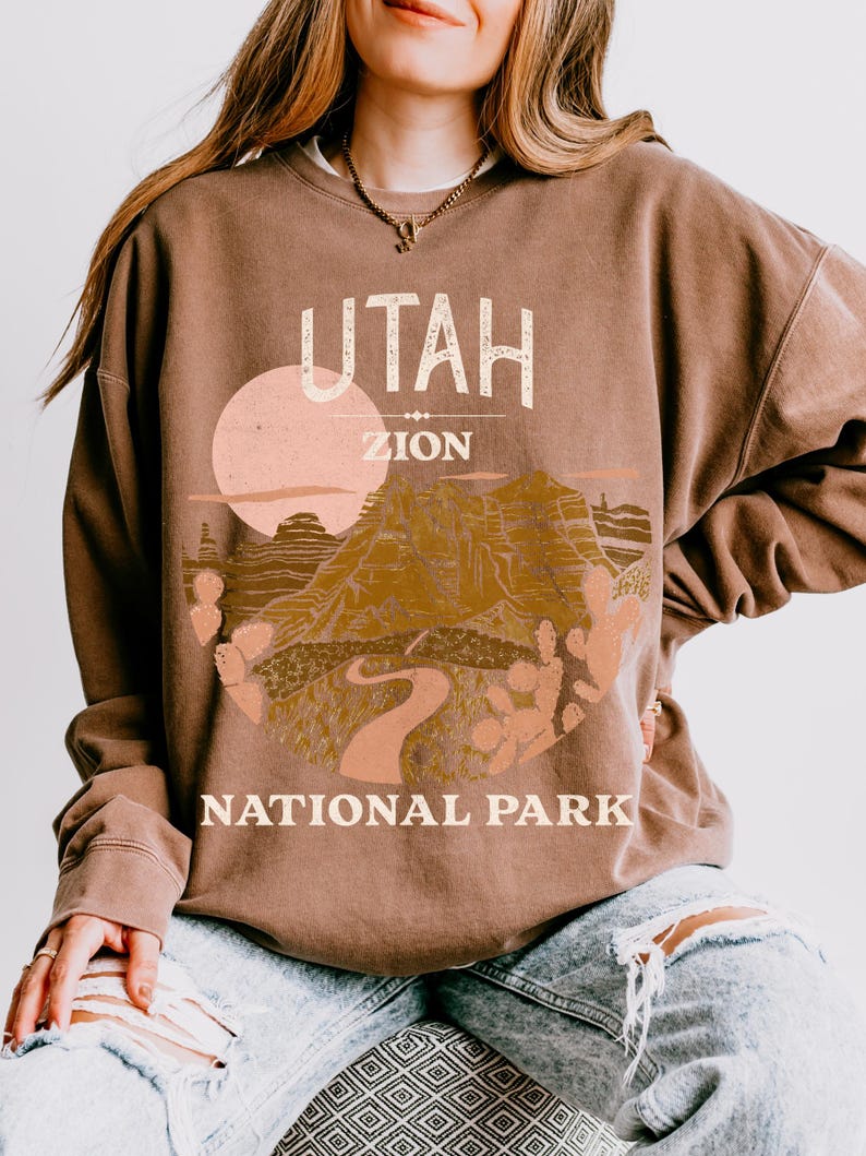 Woman wearing an Espresso oversized sweatshirt with a vintage graphic of Zion National Park in Utah, featuring canyons, cacti, and a sunset design, styled with ripped jeans.