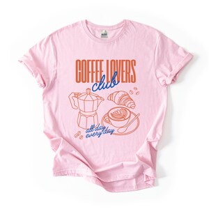 A  flat lay of a Pink Gildan 2000 t-shirt with a design featuring a hand-drawn coffee and croissant design that reads Coffee Lover Club, All Day Every Day.