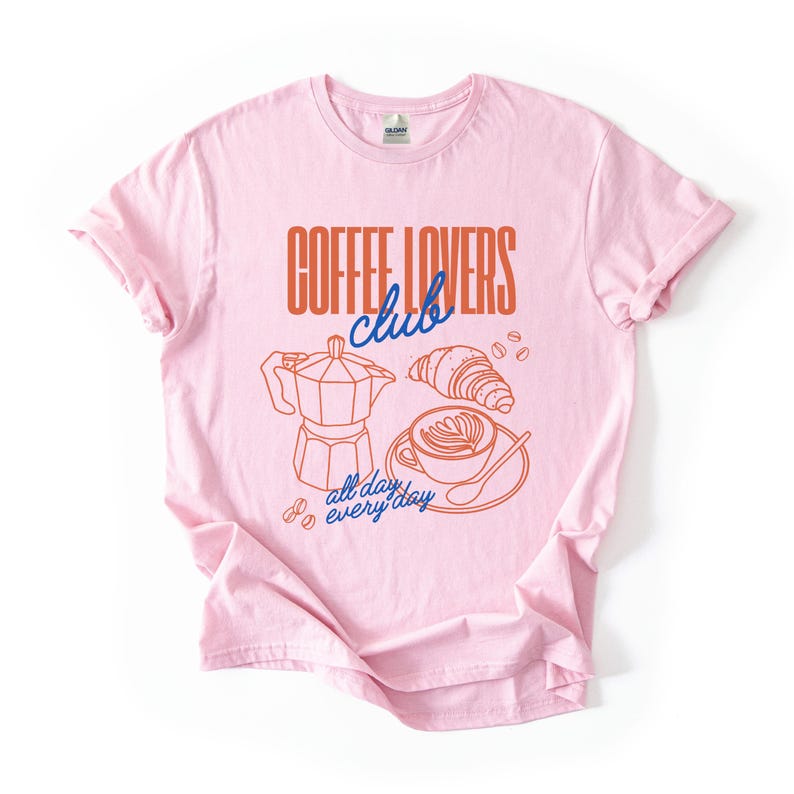 A  flat lay of a Pink Gildan 2000 t-shirt with a design featuring a hand-drawn coffee and croissant design that reads Coffee Lover Club, All Day Every Day.