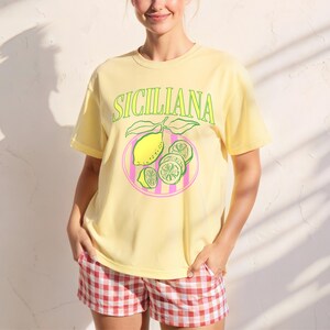 A woman is wearing a Banana Comfort Colors shirt with a design featuring a hand-drawn lemon and lemon slices on a plate, and the phrase Siciliana at the top in a serif font.