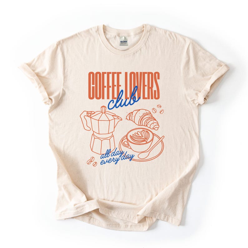 A  flat lay of a Natural Gildan 2000 t-shirt with a design featuring a hand-drawn coffee and croissant design that reads Coffee Lover Club, All Day Every Day.